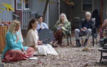 Photo of a cacao ceremony in a previous retreat. One female retreat leader is playing a drum and the other sits beside her. They are outdoors, surrounding a fire pit with retreat participants.