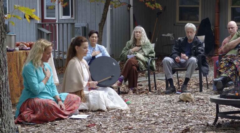Photo of a cacao ceremony in a previous retreat. One female retreat leader is playing a drum and the other sits beside her. They are outdoors, surrounding a fire pit with retreat participants.