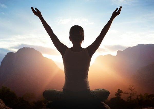 Woman sitting cross-legged with her arms outstretched above her head. She is facing the sun setting over the mountains.