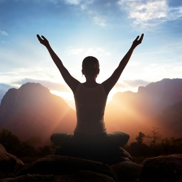 Woman sitting cross-legged with her arms outstretched above her head. She is facing the sun setting over the mountains.