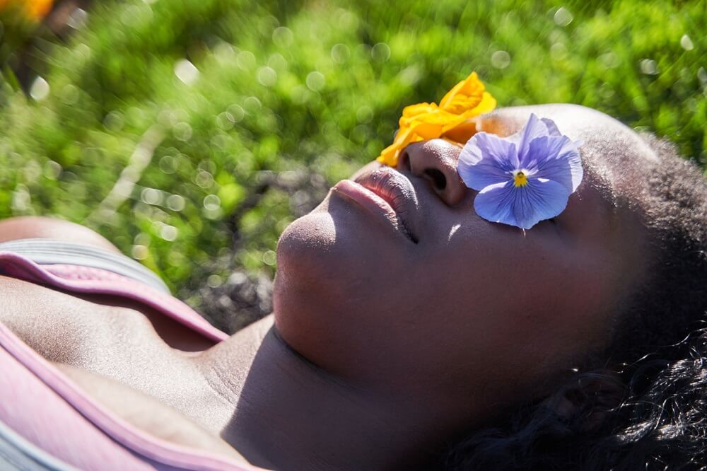 woman-lying- on-grass-with-yellow-&-purple-flower-blossoms-on-eyes