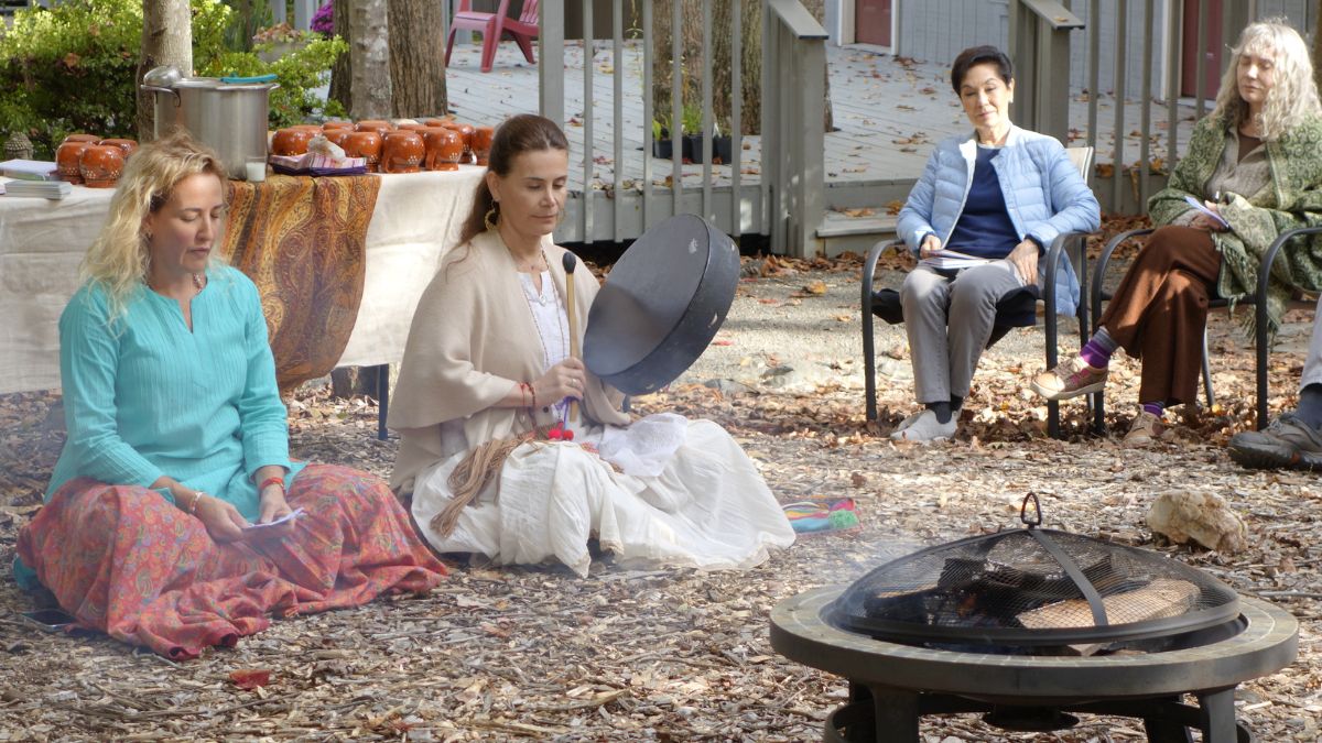 Photo of an outdoor cacao ceremony with two female retreat leaders, one is playing a drum. A couple of participants sit nearby in the circle, meditatively watching.