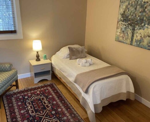 Cozy private guest room at Sanctuary Retreat Center with a twin bed, bedside lamp, upholstered chair, patterned rug, and warm natural light.