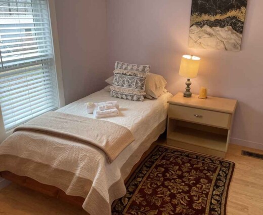 Cozy private guest room at Sanctuary Retreat Center with a twin bed, bedside table and lamp, rug, and warm natural light coming through the window.