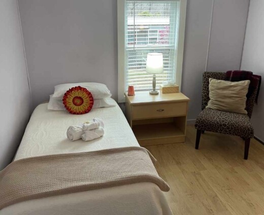 Cozy private guest room at Sanctuary Retreat Center with a twin bed, bedside table and lamp, upholstered chair, and warm natural light.