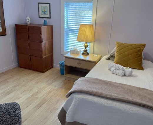 Cozy private guest room at Sanctuary Retreat Center with a twin bed, bedside table and lamp, upholstered chair, and warm natural light.