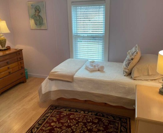 Cozy private guest room at Sanctuary Retreat Center with a twin bed, bedside table and lamp, upholstered chair, rug, chest of drawers, and warm natural light coming through the window.